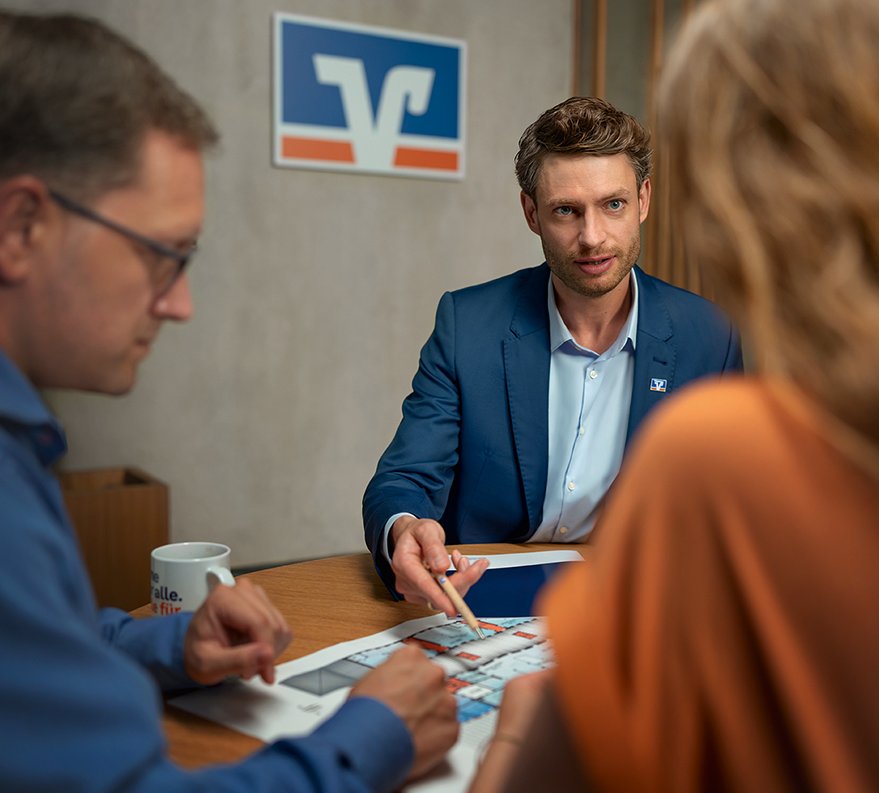 Ein Berater einer Bank zeigt auf einen Bauplan. 