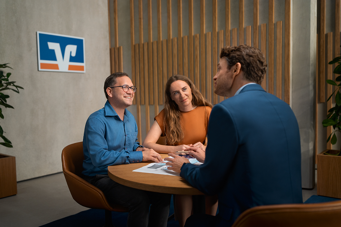Ein Mann und eine Frau sitzen an einem Tisch in einer Bankfiliale. Vor ihnen liegt ein Plan. Sie sprechen mit einem anderen Mann, ihrem Bankberater.